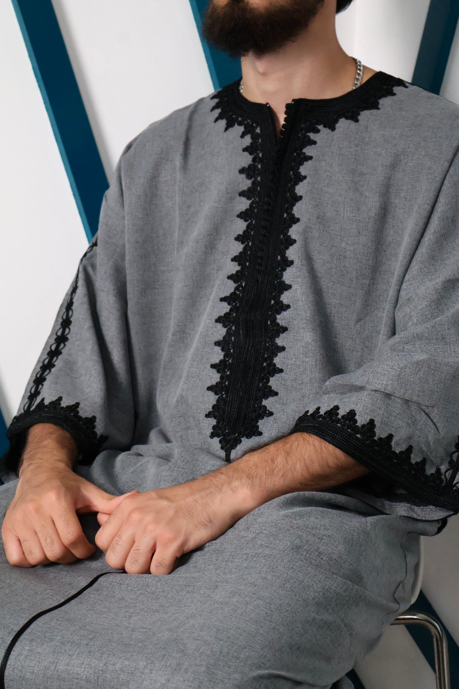 Man looking to his right, wearing a men's Moroccan Atlas gandoura with detailed black embroidery, standing against a building. #colour_grey