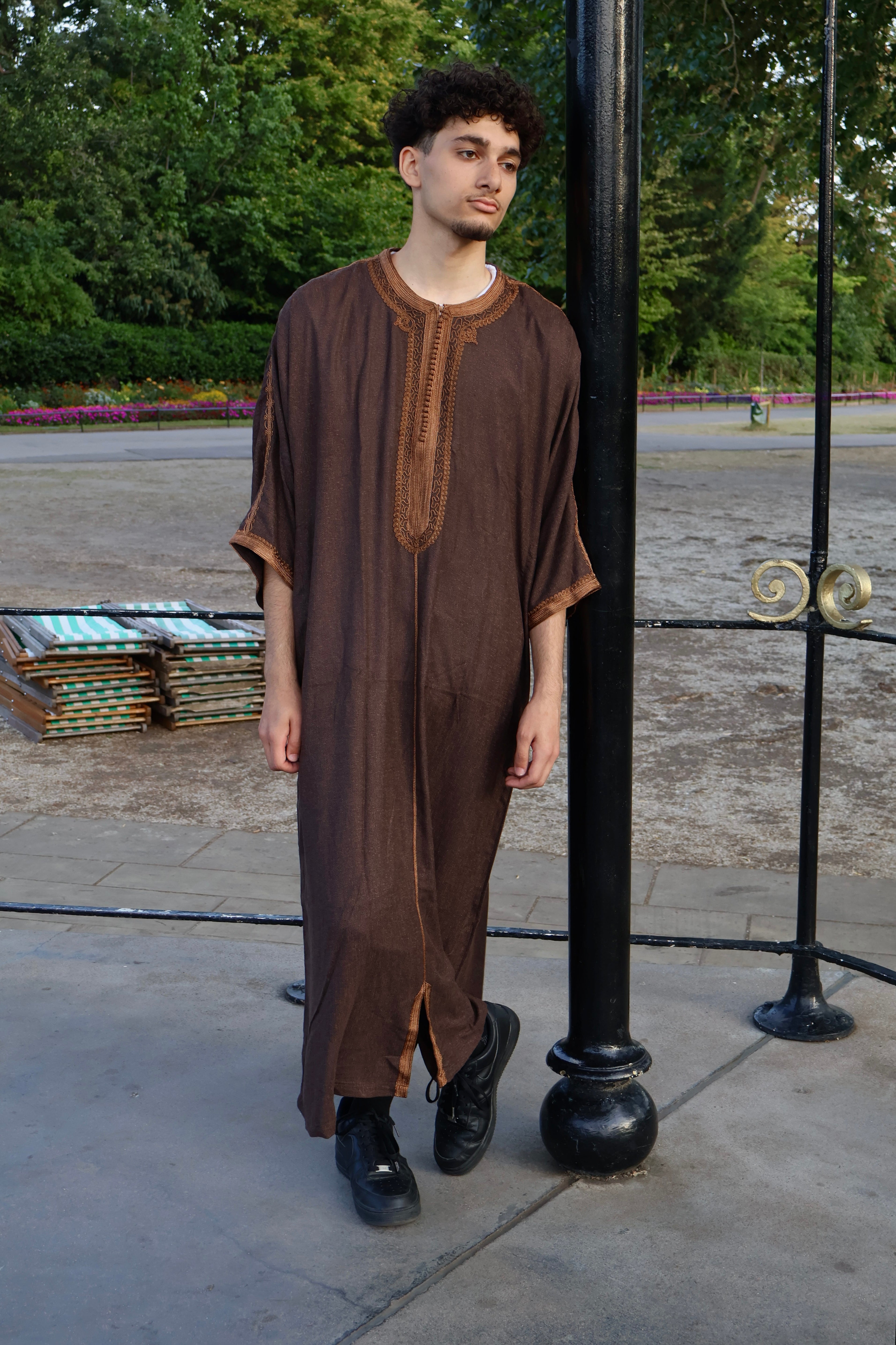 Man leaning against a black pole outdoors, wearing a rich brown men&